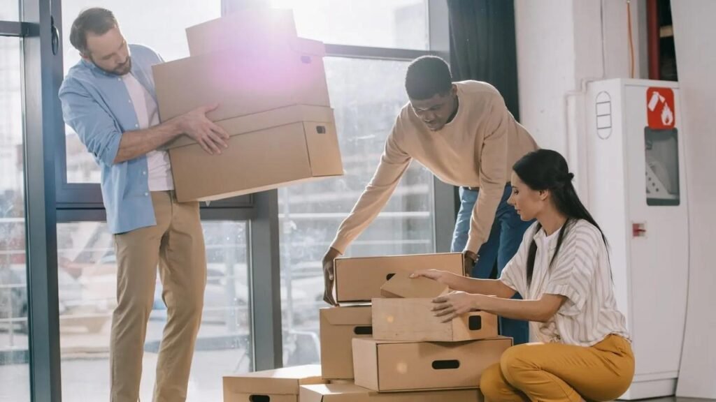 Three people are transporting boxes into a room, helping with the office moving process.
