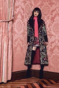 A woman wearing a pink sweater and coat poses against a pink wall, highlighting the fashion of Stacey Bendet, Alice Olivia.
